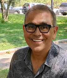 "Portrait of a man shoulders up wearing plastic rimmed glasses, a collared shirt and big smile. Background an outdoor street scene"